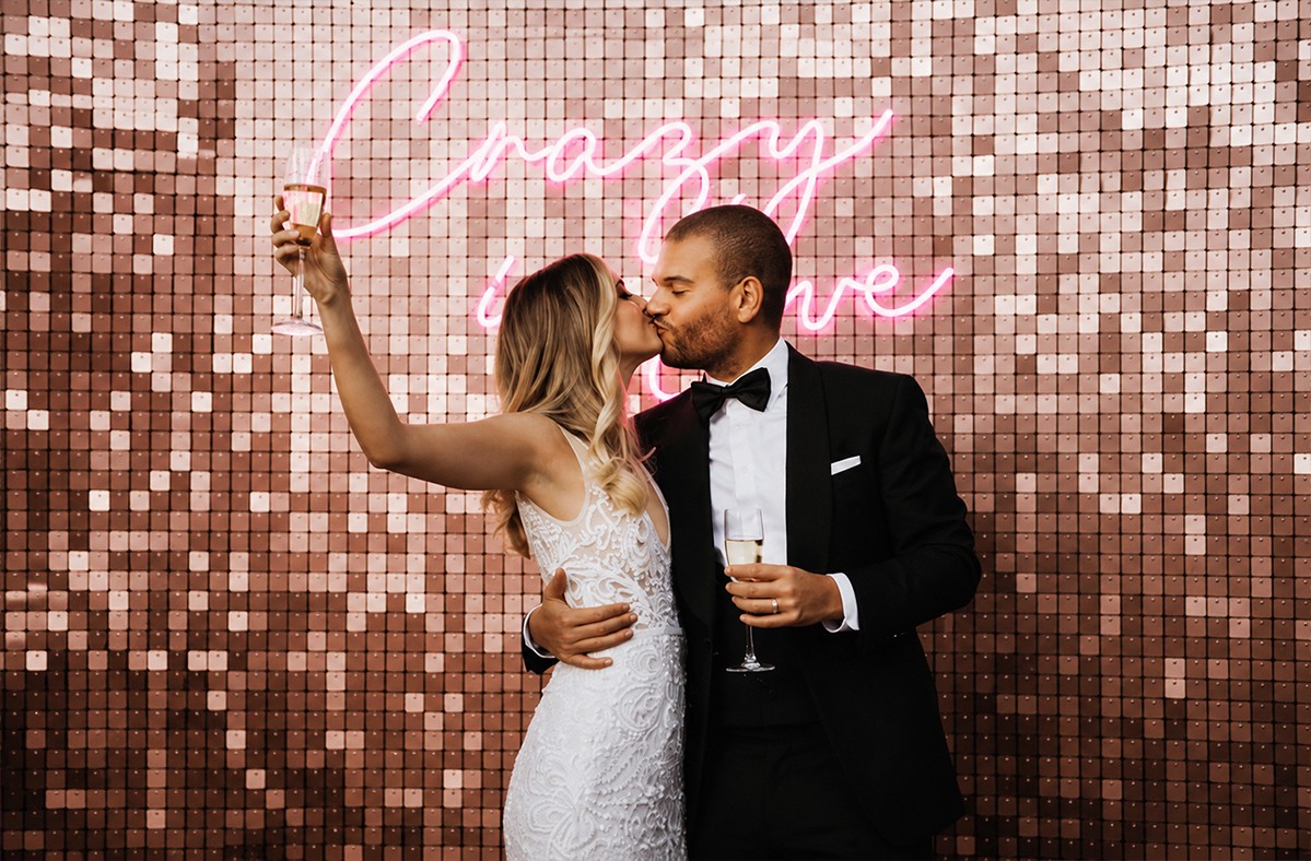 Bruidspaar kust voor een roségouden shimmerwall met roze neonbord tijdens een feestelijk huwelijksmoment.