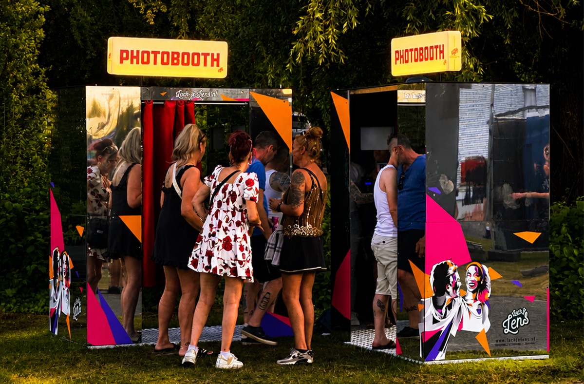 Bezoekers wachten bij twee spiegel-photobooths op een festival, terwijl een groep vrienden zich klaarmaakt voor een fotosessie.