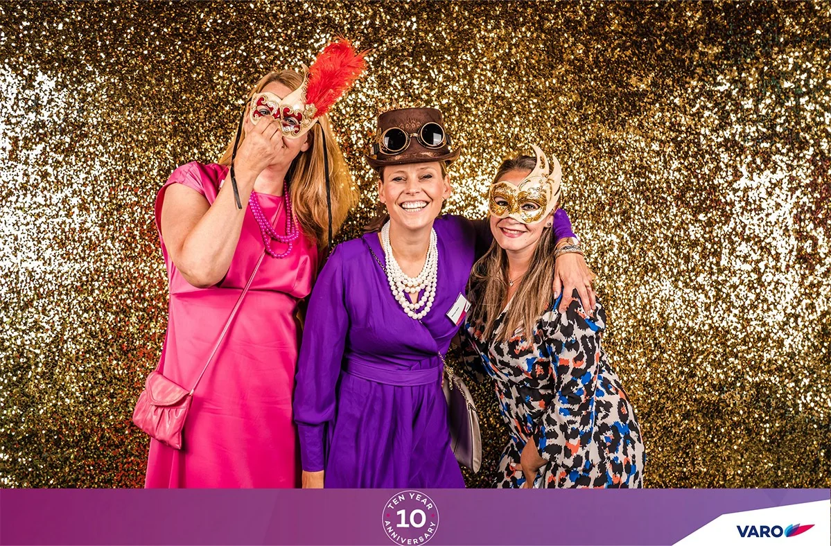 Drie vrouwen poseren verkleed bij een photobooth met gouden glitterachterwand tijdens een feestelijk evenement.
